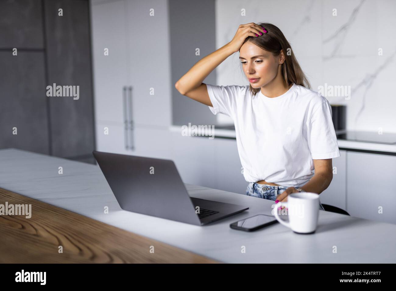 Shocked young woman sitting in the kitchen and working on laptop and counting spendings and taxes Stock Photo