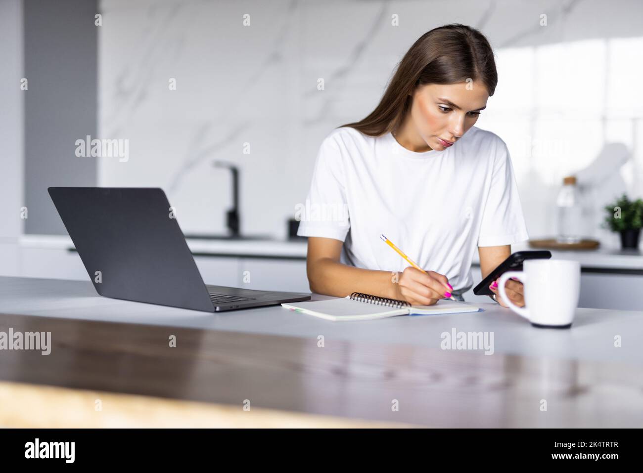 Work at home. Young woman use a laptop to work in the kitchen writes something in a notebook with a pen and speaks on the phone Stock Photo
