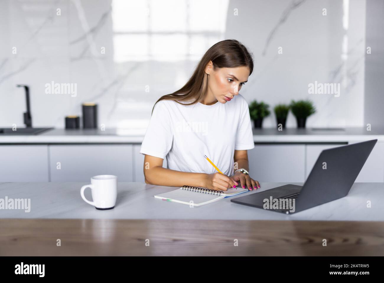 Freelance woman talks on the smartphone and takes notes with pen, using laptop for remote work from home, write down some information sitting at the t Stock Photo