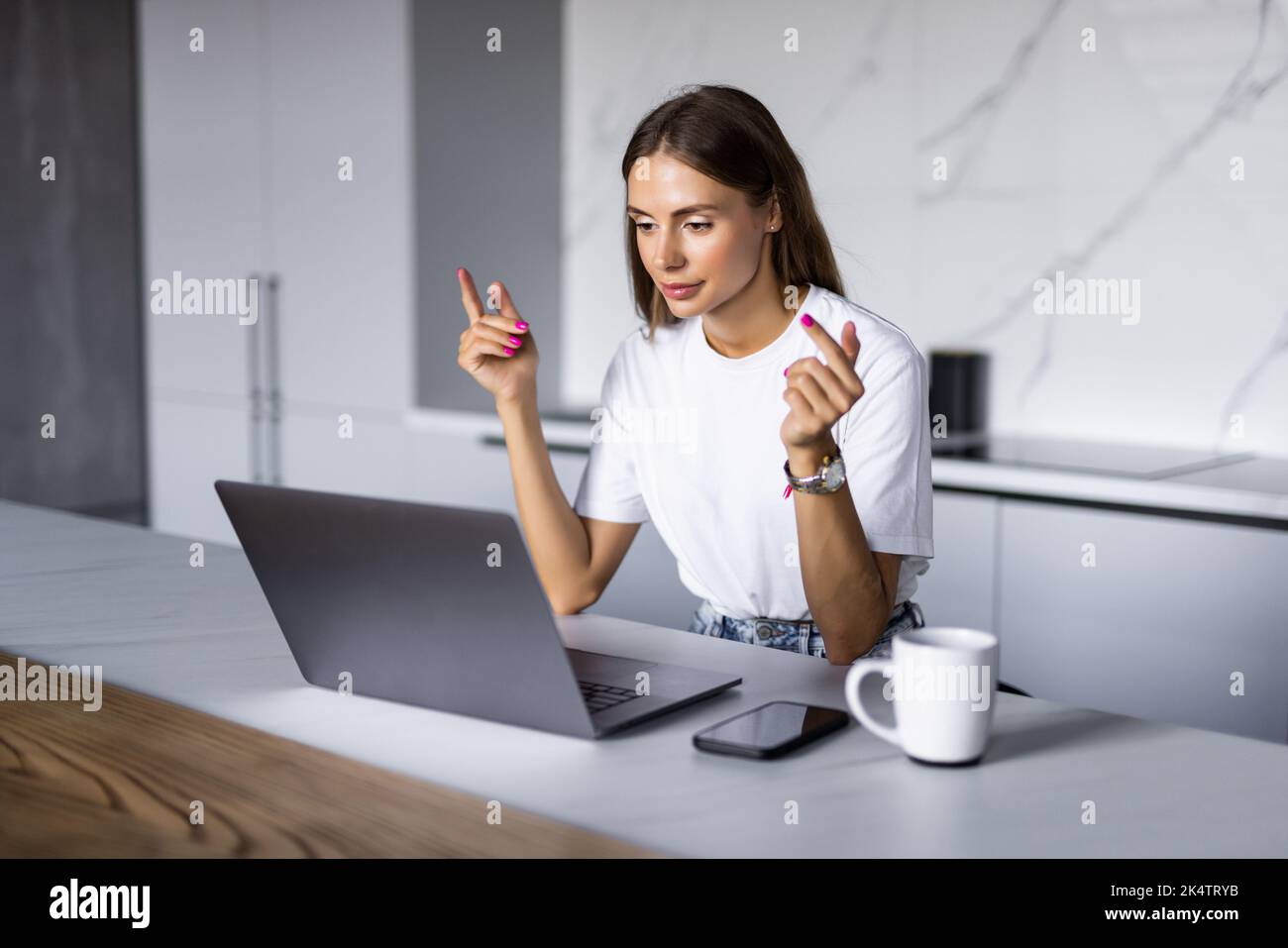 Young woman talking on video call and waving hand while sitting at table inthe kitchen. Stock Photo
