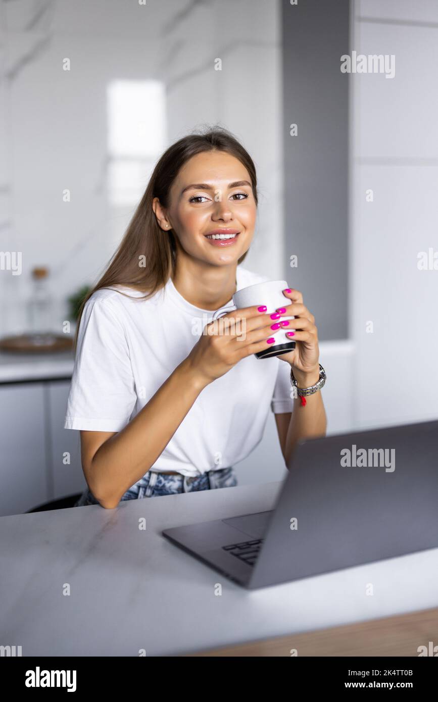 Young woman drinking morning coffee and looking at monitor of laptop while sitting in the kitchen at home Stock Photo