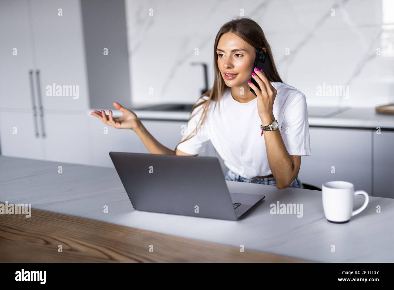 Photo of a young cheerful happy woman indoors at home at the kitchen using laptop computer talking by mobile phone. Stock Photo