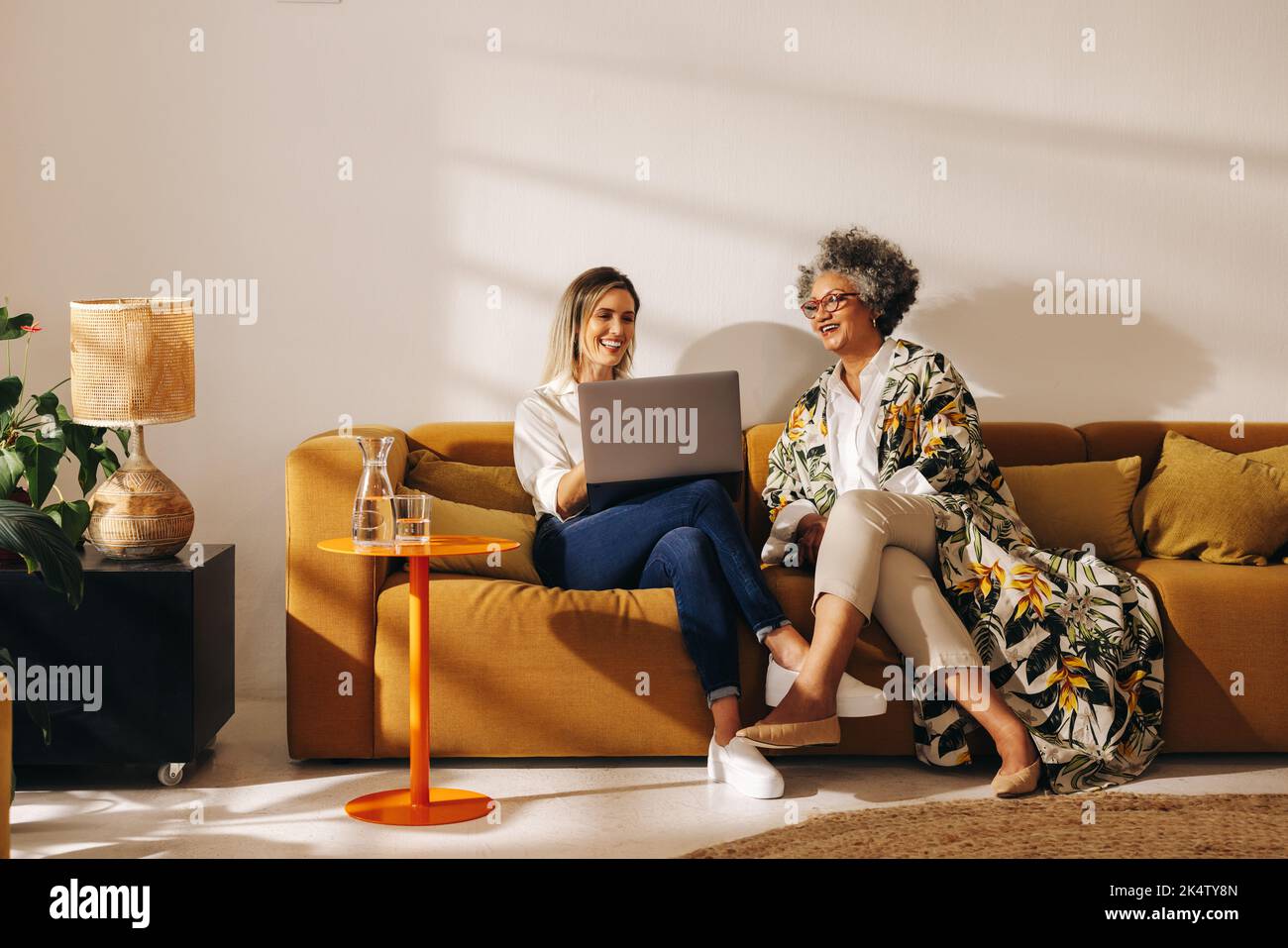 Two happy businesswomen using a laptop while working together in an office lobby. Successful female entrepreneurs sitting on a couch in a woman-owned Stock Photo