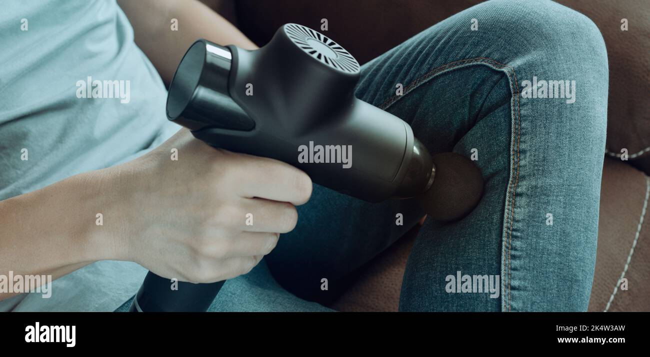 a young caucasian man, in jeans, uses a massage gun to massage the muscles of his leg, sitting on a couch, in a panoramic format to use as web banner Stock Photo