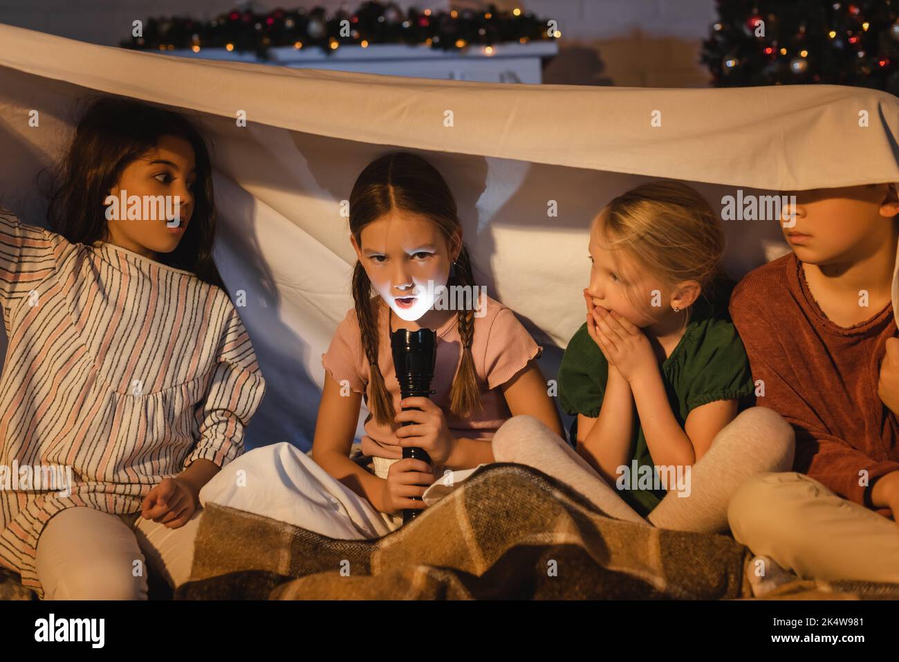 Girl holding flashlight near scared friends under blanket during christmas at home,stock image Stock Photo