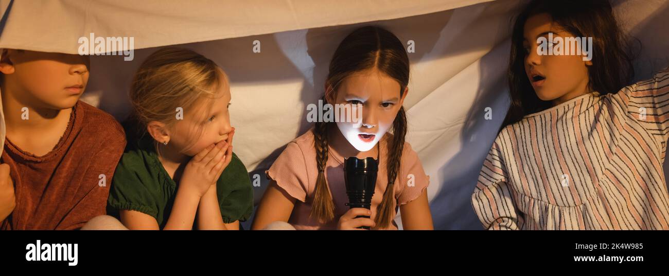 Girl holding flashlight near frightened friends under blanket at home, banner,stock image Stock Photo