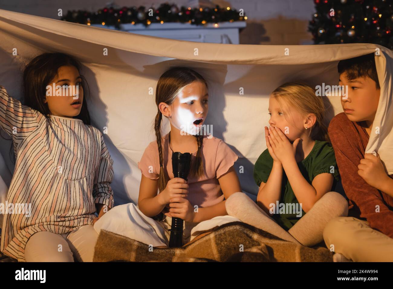 Girl holding flashlight near scared interracial friends under blanket and christmas decor at home,stock image Stock Photo