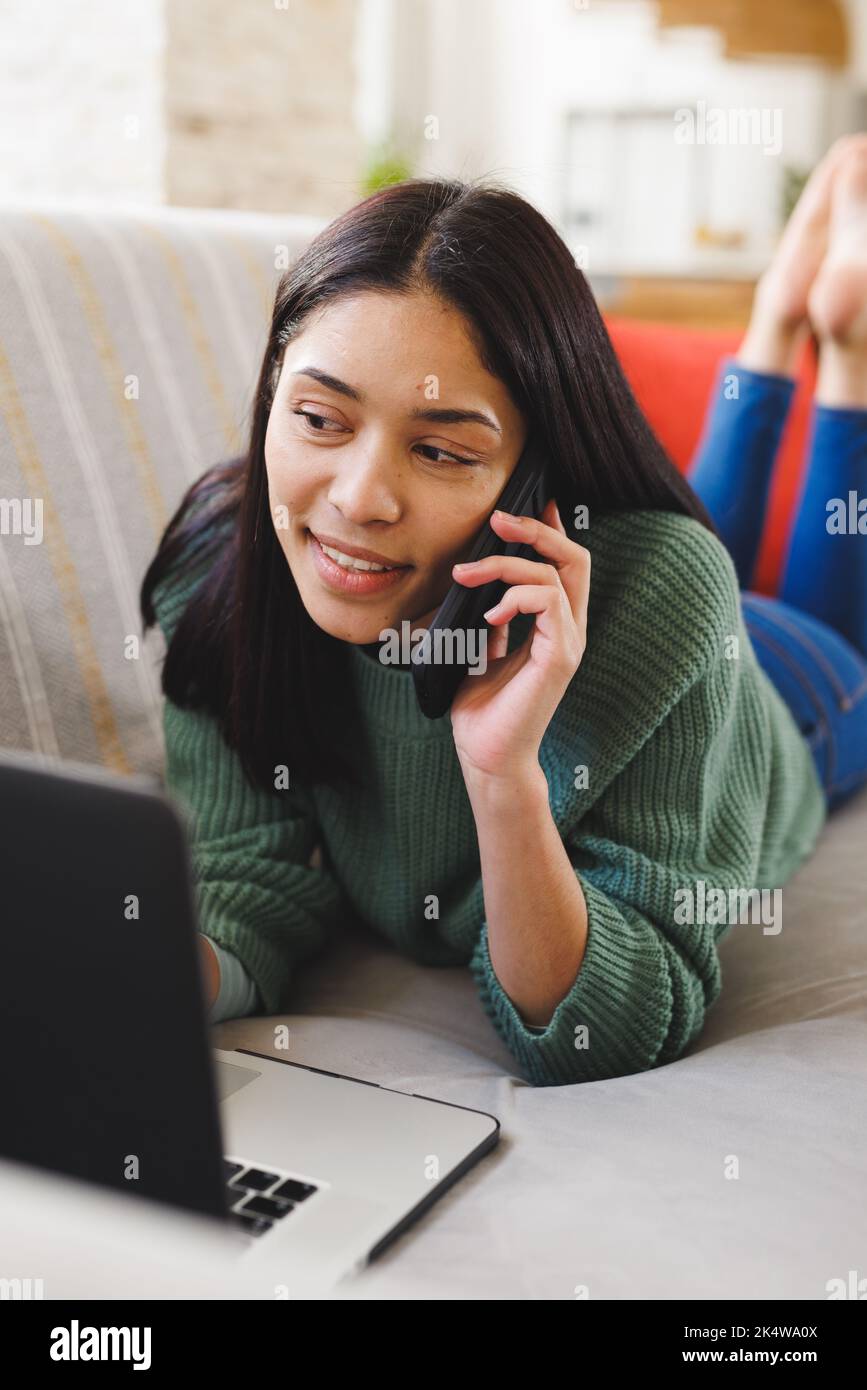 Happy biracial woman laying on sofa in living room, using laptop and ...