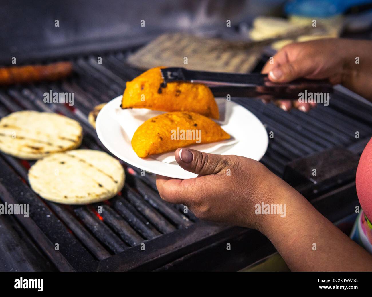 Hands serving two empanadas on a plate next to the grill with arepas