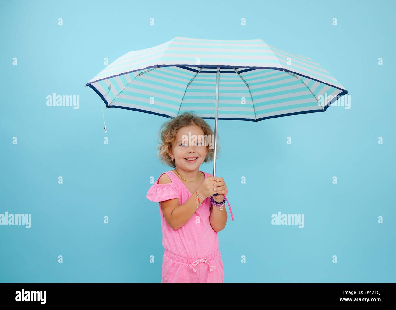Portrait of little girl with curly fair hair wearing pink jumpsuit, holding open striped umbrella on blue background. Stock Photo