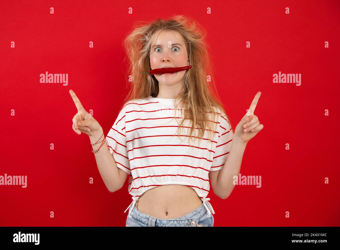 Portrait of astonished teenage girl with disheveled fair hair raising hands with fingers up keeping in mouth red chilli. Stock Photo