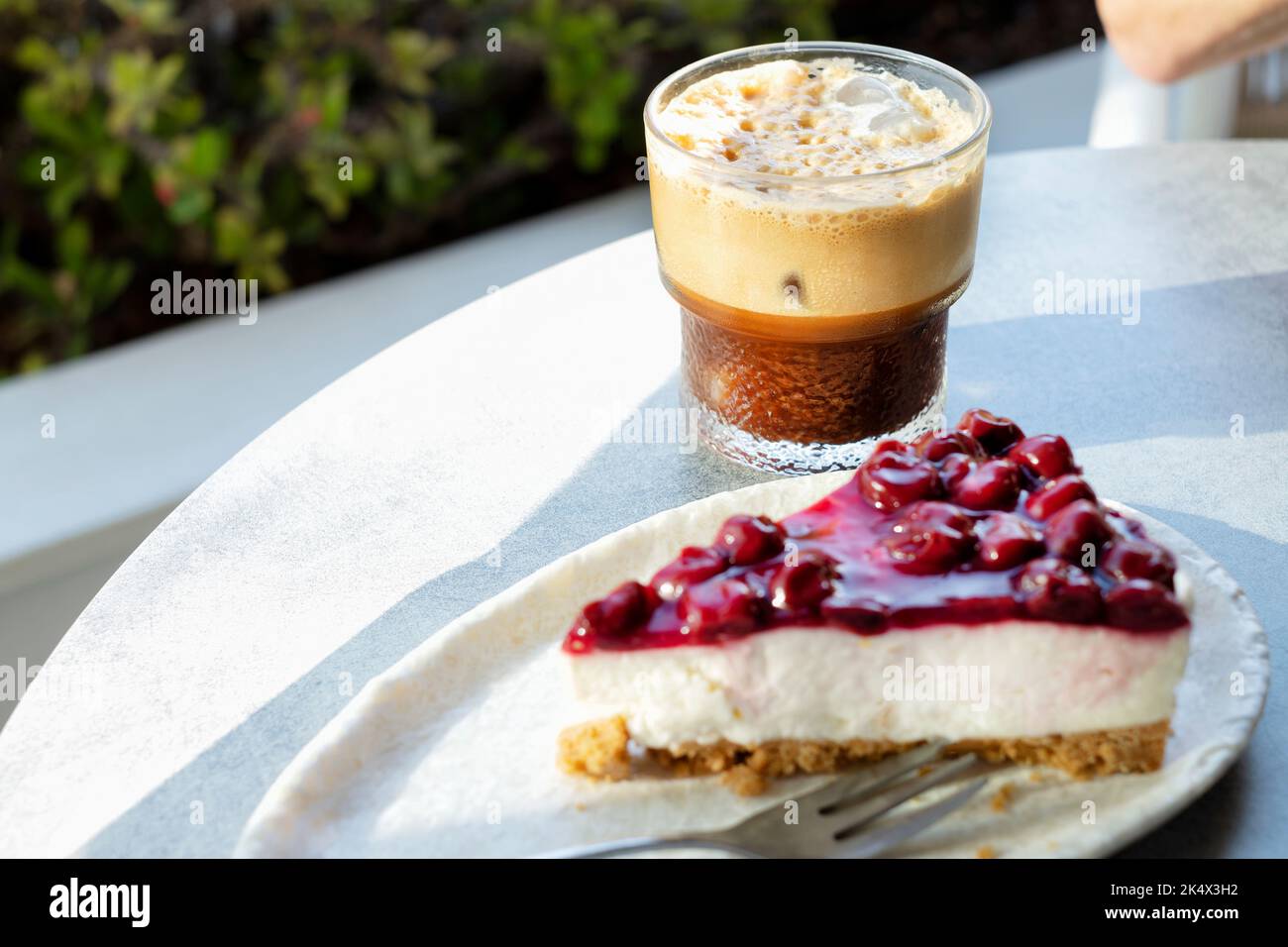 A single slice, or serving, of freshly made, indulgent, cherry cheese cake. Its served on a plate with an iced coffee to an outside table in a cafe. Stock Photo