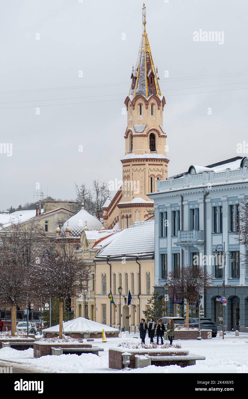 Beautiful street in Rotuse square, Vilnius Old Town, with girls walking, Saint Nicolas church, old buildings and street lamps in winter with snow Stock Photo