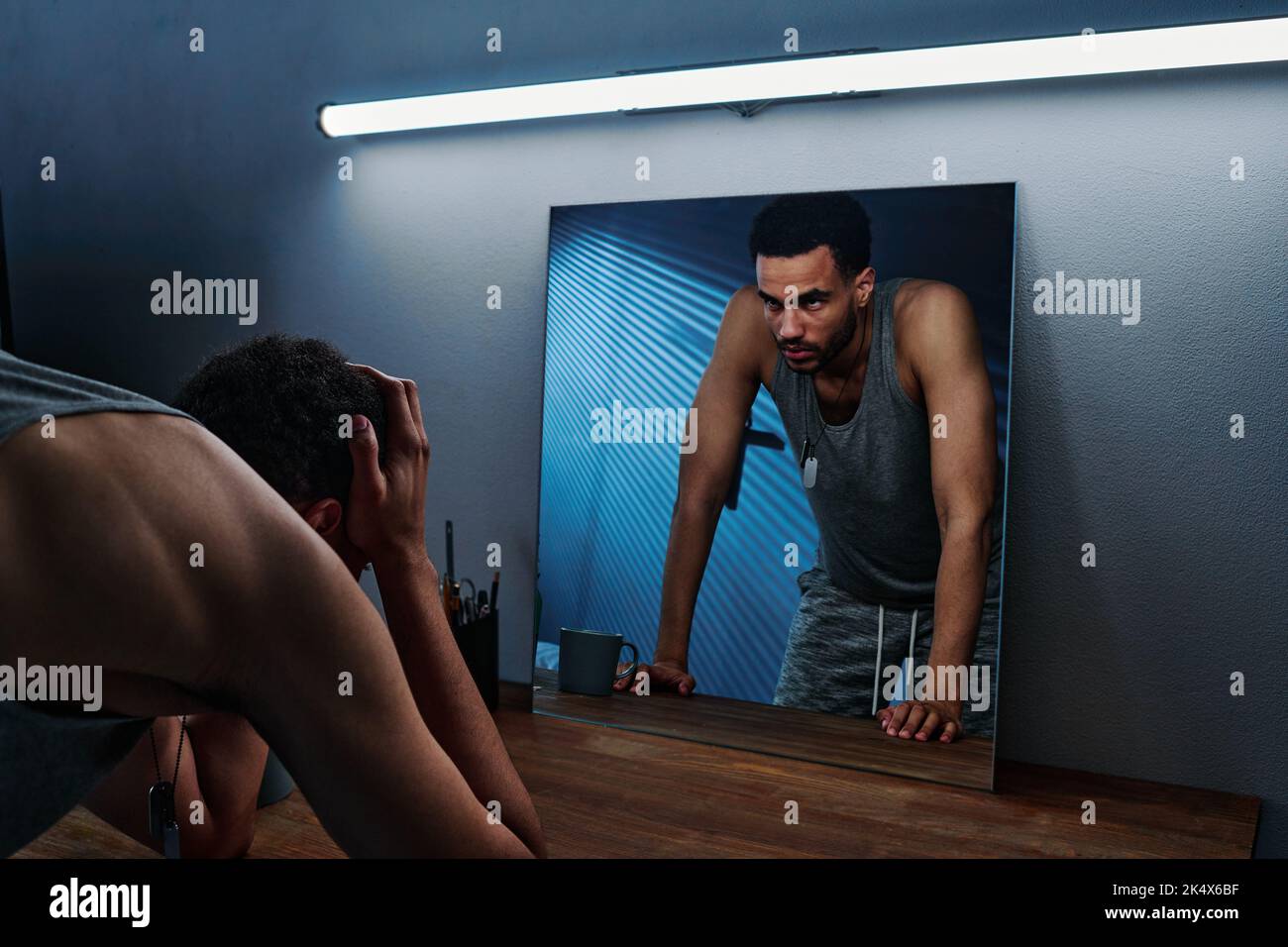 Young insomniac man holding head in hands in front of mirror with reflection of him expressing anger and aggression Stock Photo