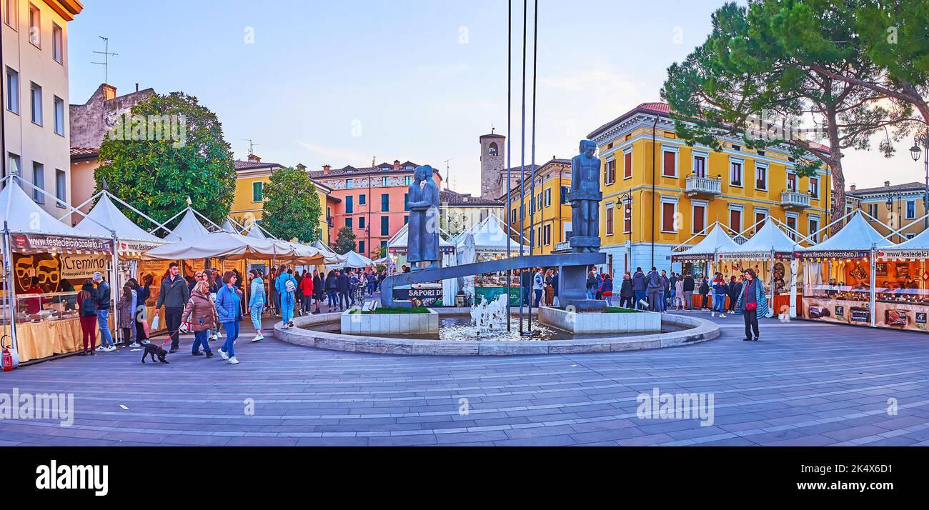 DESENZANO DEL GARDA, ITALY - APRIL 10, 2022: The evening on Piazza Cappelletti square with market stalls, statue by Mario Gatti and houses in backgrou Stock Photo