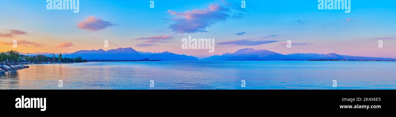 The purple and blue silhouettes of Garda Prealps behind the Lake Garda, reflecting sunset sky with pink clouds, Desenzano del Garda, Italy Stock Photo