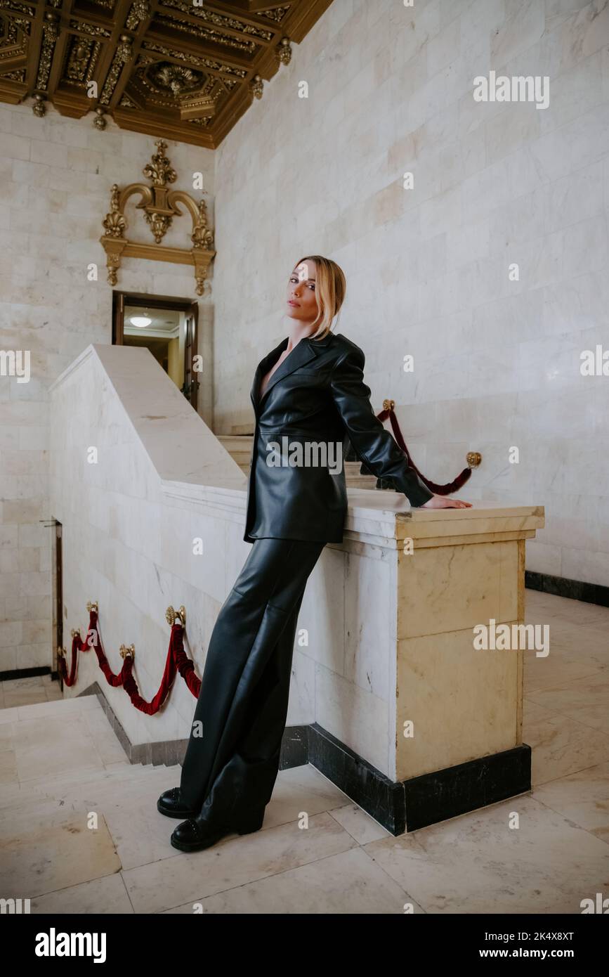 Pretty slim lady wearing a dark leather suit standing in a majestic, historic interior, leaning against a marble wall Stock Photo