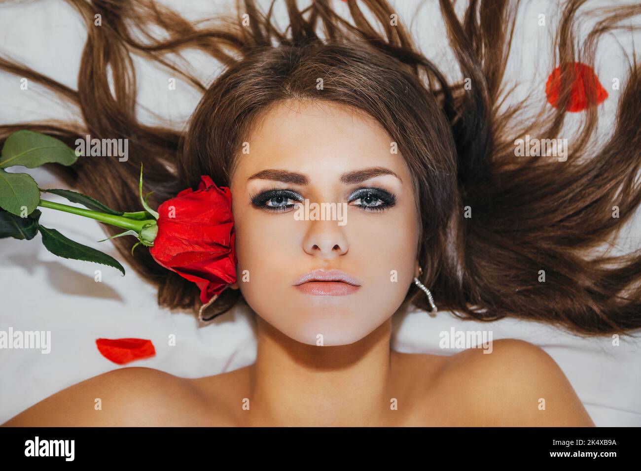 Portrait (taken from above) of sensual brunette girl on a white sheet with rose petals, holding a red rose near her face; her hair is spread out Stock Photo