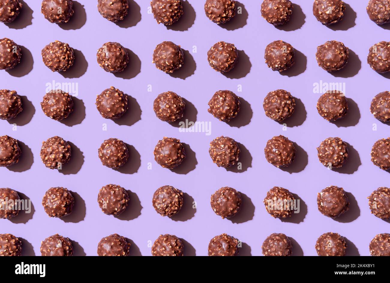 Full-frame background with many chocolate truffles aligned symmetrically on a purple table. Chocolate and hazelnuts candies in bright light on a vibra Stock Photo