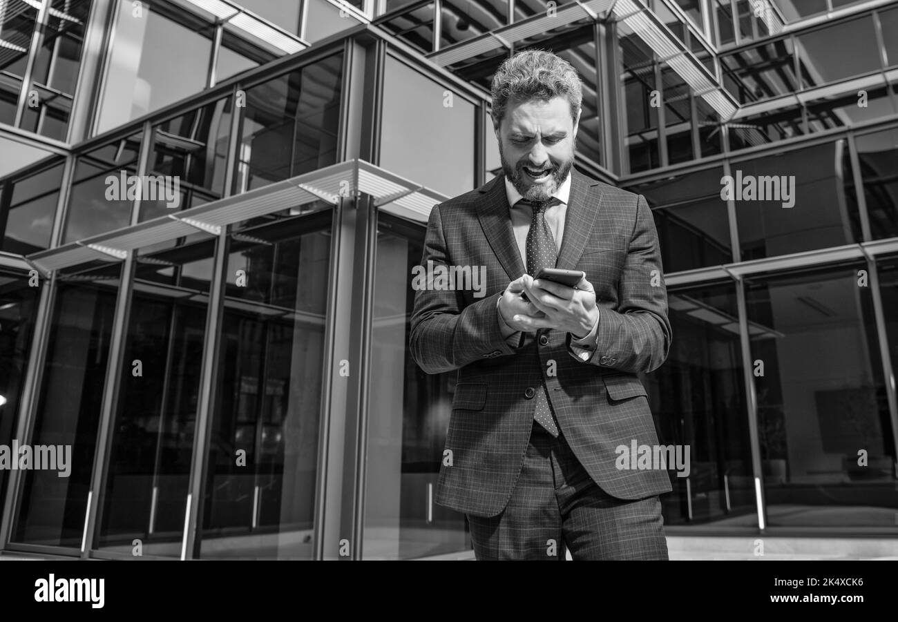 confused mature business man in formalwear holding phone outdoor, panic Stock Photo