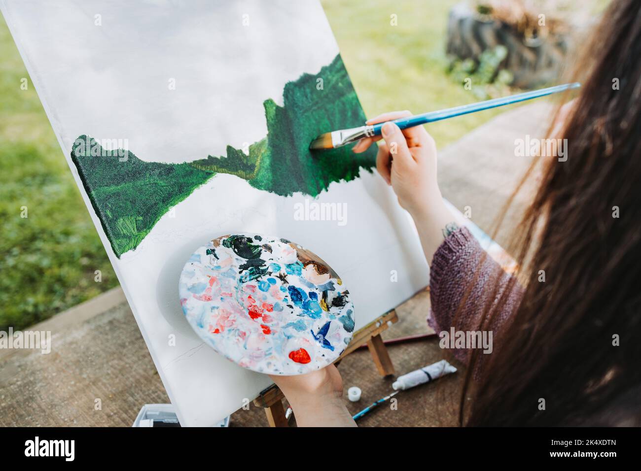 Unrecognizable young female artist painting her own mountain scenery with oil on a canvas. Amateur painter. Stock Photo