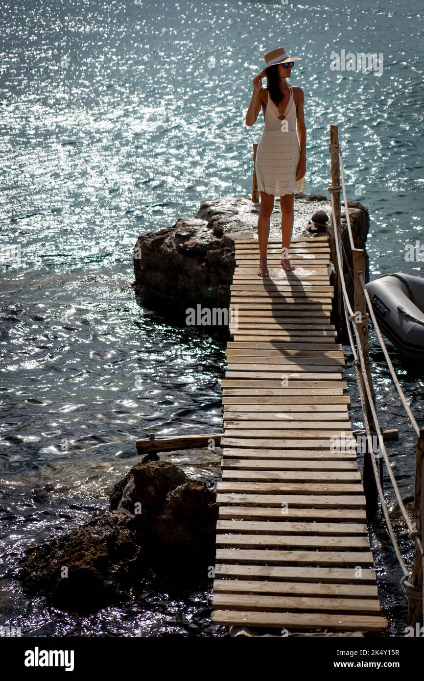 Brunette woman with hat and light beige dress walking on walkway over the sea Stock Photo