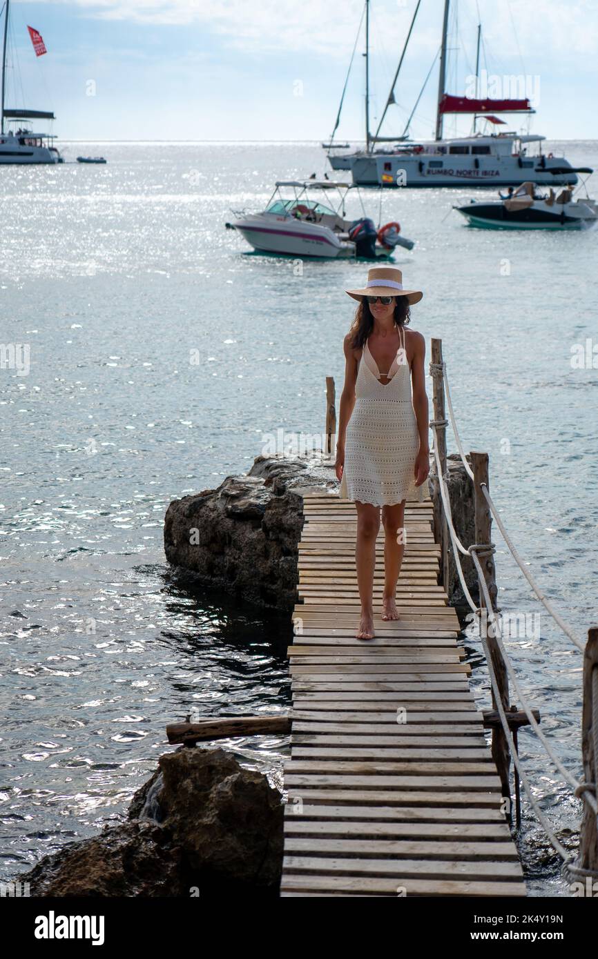 Brunette woman with hat and light beige dress walking on walkway over the sea Stock Photo