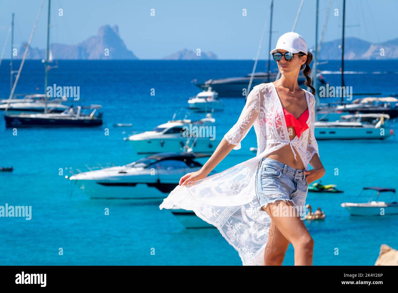 Brunette woman with white cap, crochet dress and shorts posing next to yachts in Formentera Stock Photo