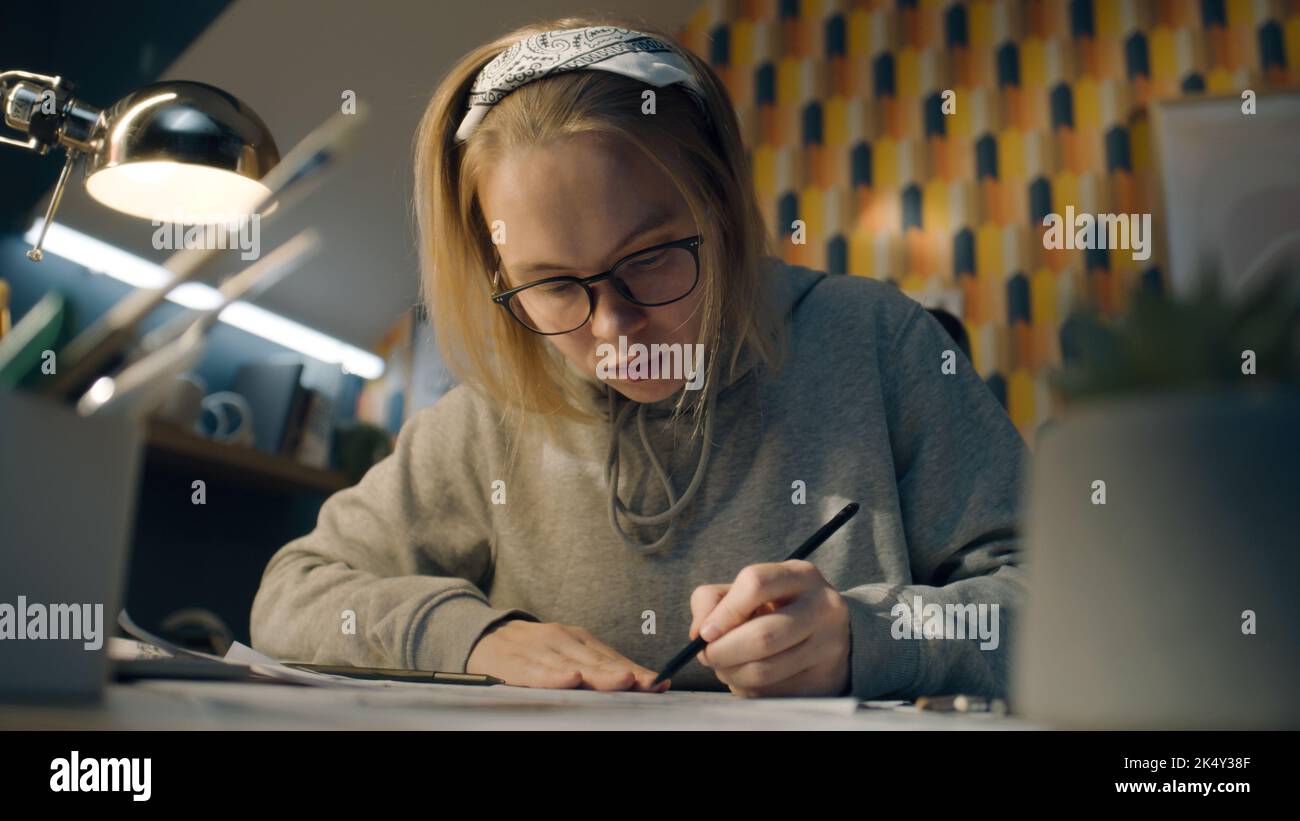 Focused illustrator draws sketches for her project. Young female artist with glasses dressed in a grey hoodie and sits on her work desk. Makes pencil sketches. Story telling concept. Stock Photo