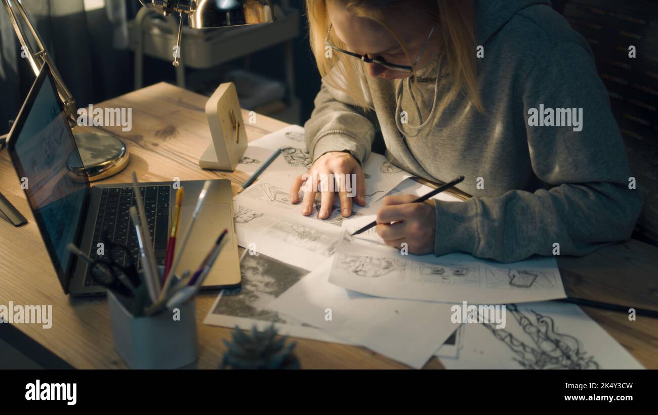 Young creative designer walks towards her work desk in a home based studio. The woman sits on the chair, puts on her glasses and starts sketching with a pencil. Stock Photo