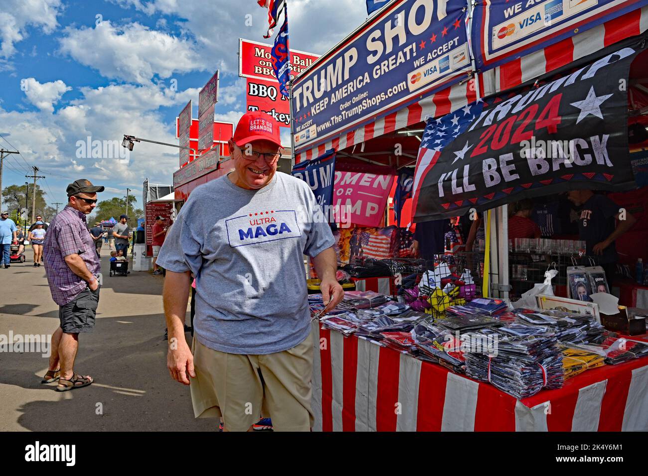 HUTCHINSON, KANSAS - SEPTEMBER 10, 2022 Man wearing a Ultra MAGA t-shirt and a red Let’s go Brandon hat poses in front of the booth  selling Trump flags, souvenirs and other items at the Kansas State Fair HUTCHINSON, KANSAS - SEPTEMBER 10, 2022HUTCHINSON, KANSAS - SEPTEMBER 10, 2022 Stock Photo