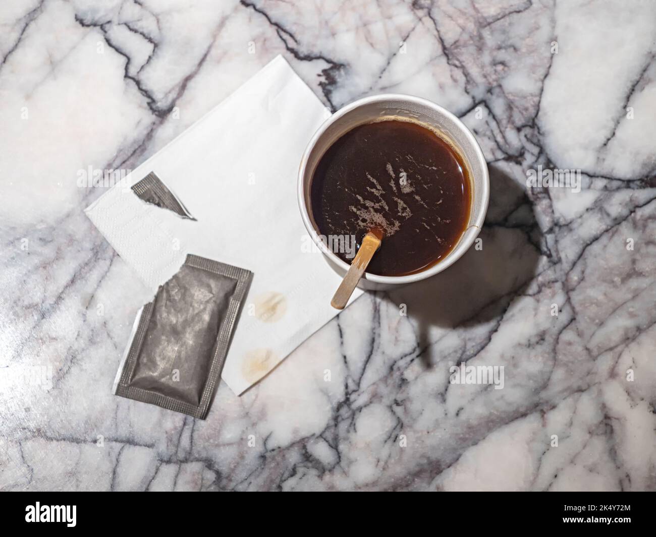 Black coffee in the brown disposable cup, a torn bag of  sugar and the wooden stirring stick on the marble table top background Stock Photo