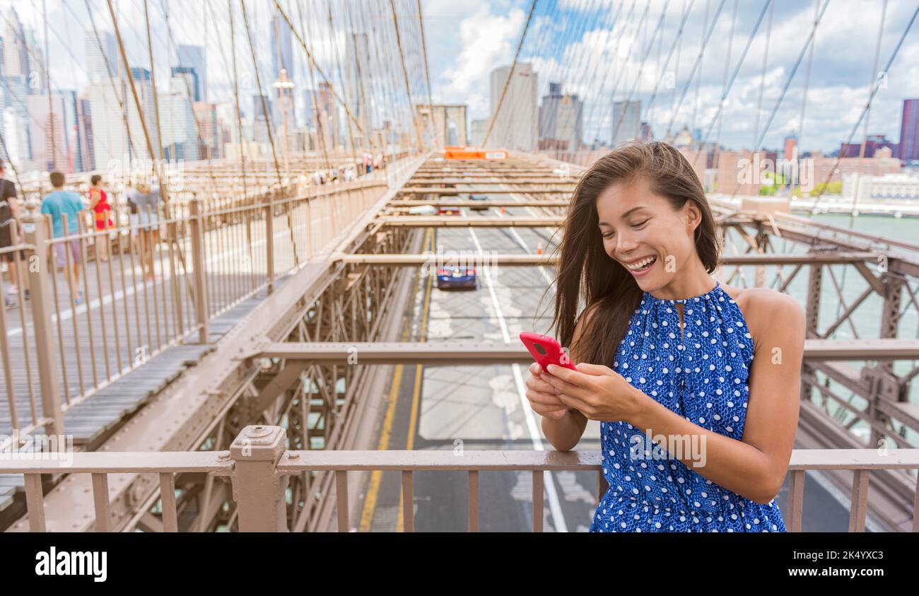 New York woman using phone app walking on Brooklyn Bridge towards Manhattan city skyline. Young female professional multicultural lady, New York City Stock Photo