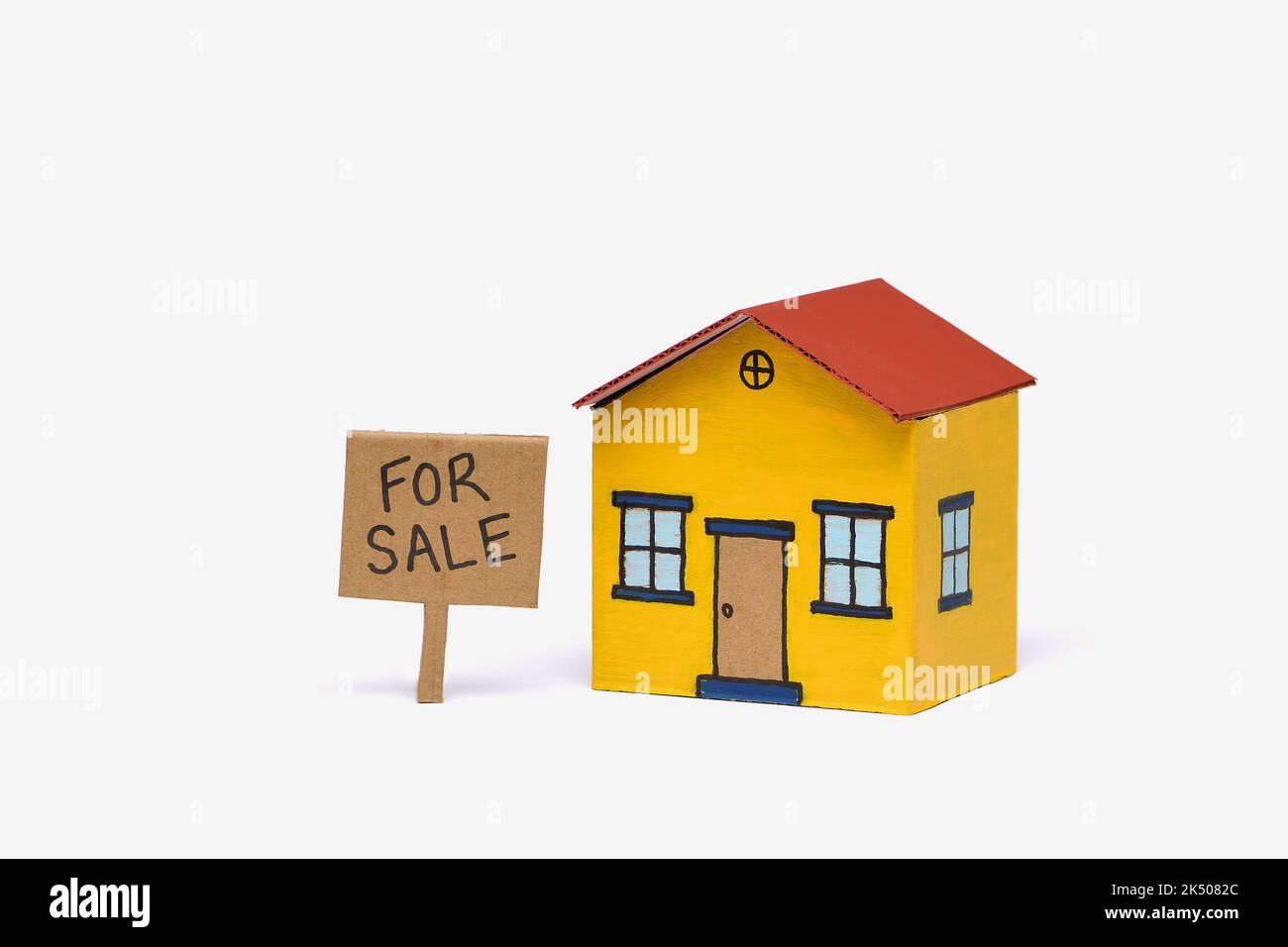 A yellow and red toy cardboard house in the middle of frame isolated on a white background with a Realestate For Sale sign, captured in a studio Stock Photo