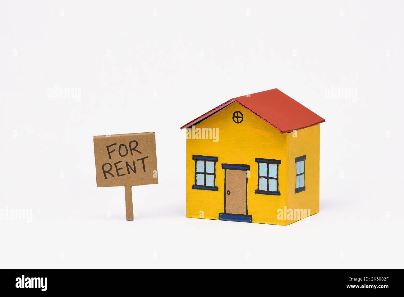 A yellow and red toy cardboard house in the middle of frame isolated on a white background with a Realestate For Rent sign, captured in a studio Stock Photo