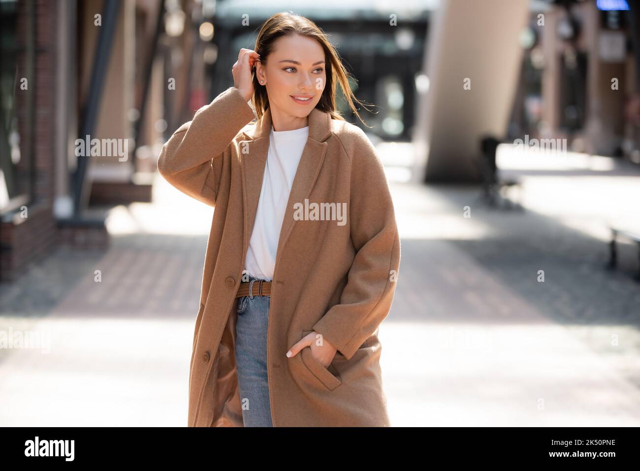 young woman standing with hand in pocket of beige coat and looking away on blurred street,stock image Stock Photo