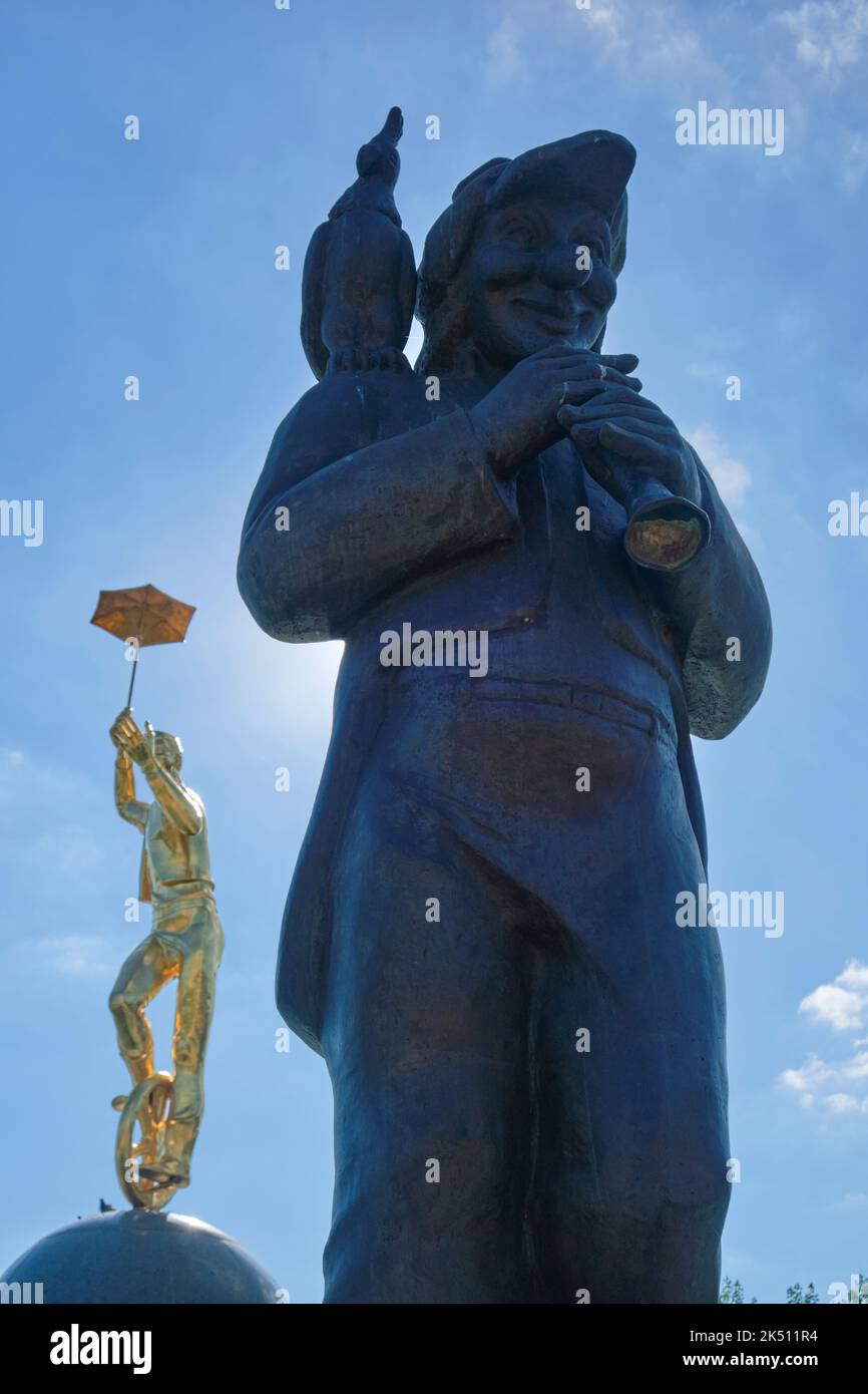 A bronze sculpture of a pirate with a parrot on his shoulder. A gold sculpture of a boy riding a unicycle is in the background.  In the plaza in front Stock Photo