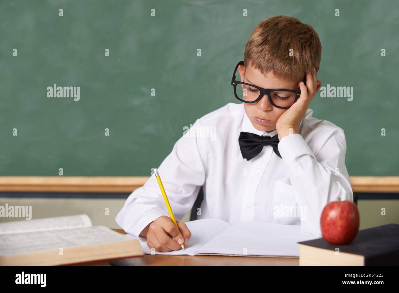 Not another test. A young boy with glasses and a bow-tie sitting in class writing despondently in a book. Stock Photo