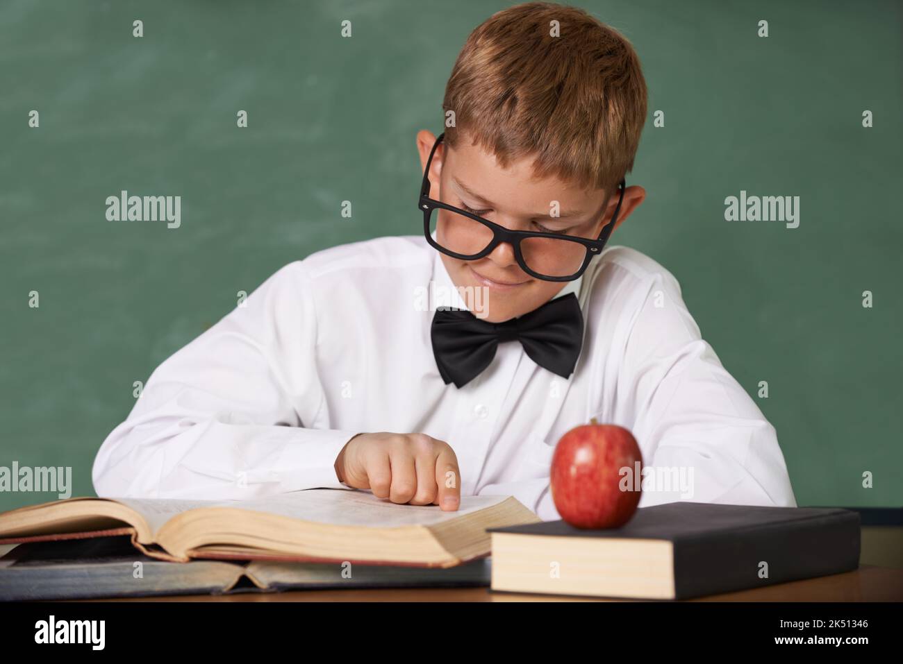 He loves to read. A young boy wearing glasses and a bow-tie concentrating on his reading with a red apple on the table in front of him. Stock Photo