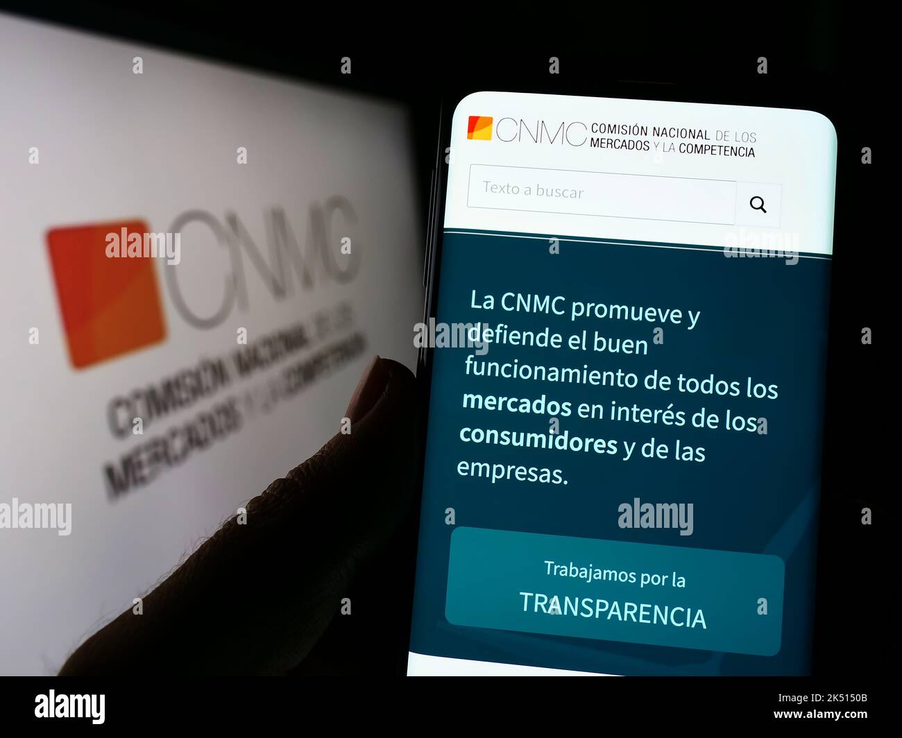 Person holding cellphone with webpage of Spanish competition regulator CNMC on screen in front of logo. Focus on center of phone display. Stock Photo