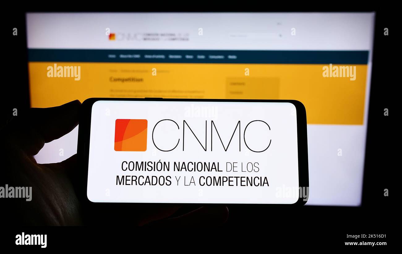 Person holding mobile phone with logo of Spanish competition regulator CNMC on screen in front of web page. Focus on phone display. Stock Photo
