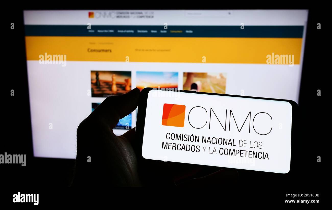 Person holding cellphone with logo of Spanish competition regulator CNMC on screen in front of webpage. Focus on phone display. Stock Photo