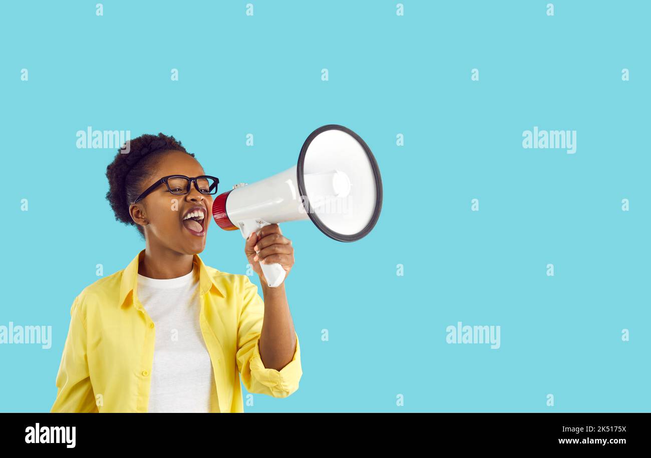 Energetic young African American woman shouting into megaphone standing on turquoise background Stock Photo
