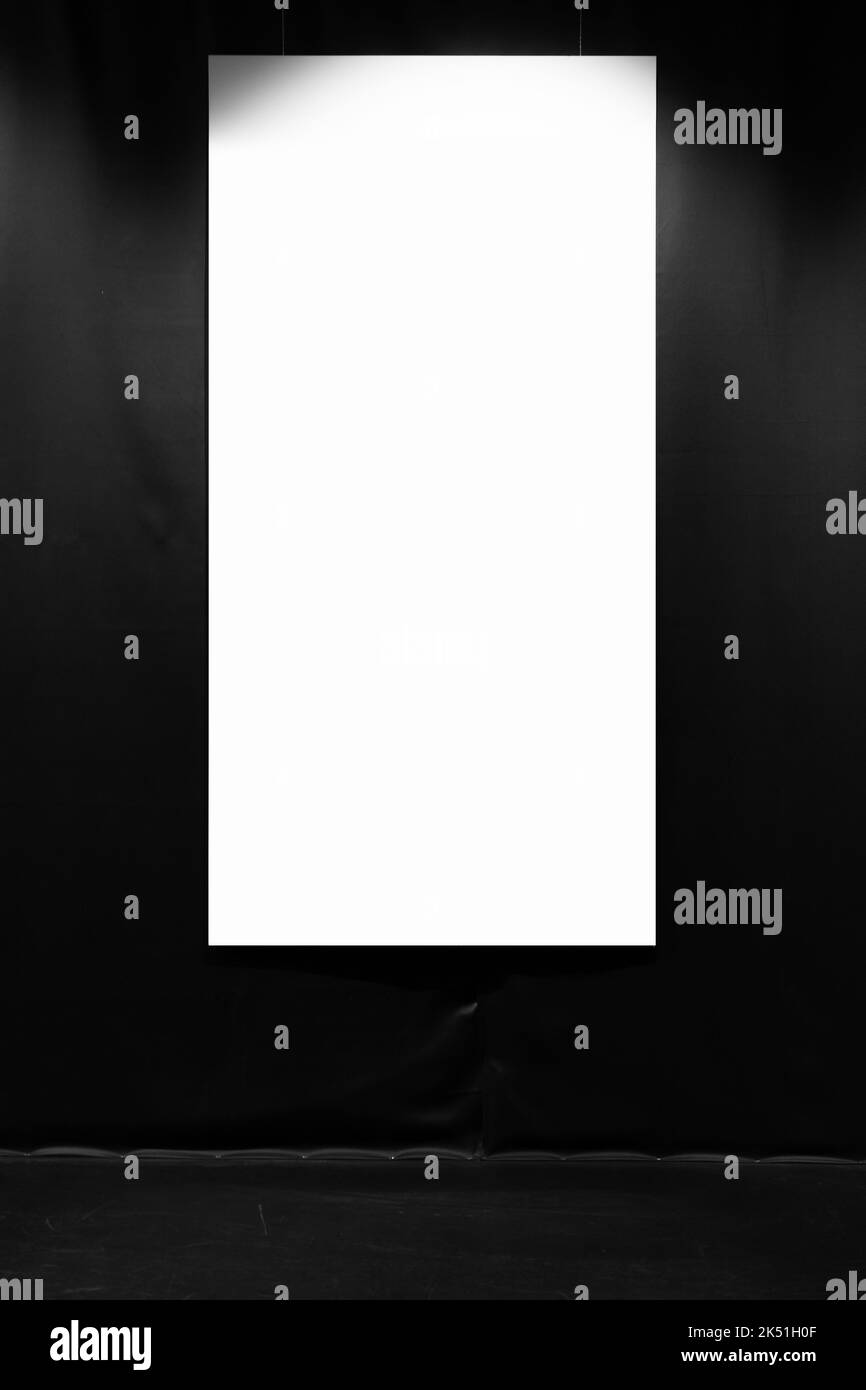 Blank white vertical poster with copy space area hangs on a black wall in a dark exhibition hall. Front view, background photo Stock Photo