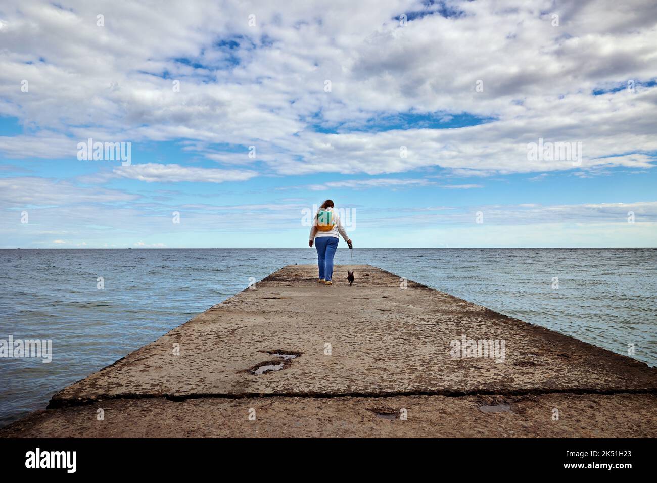 A woman with a small dog on a leash walk along the pier towards the blue sky with white cirrus clouds Stock Photo