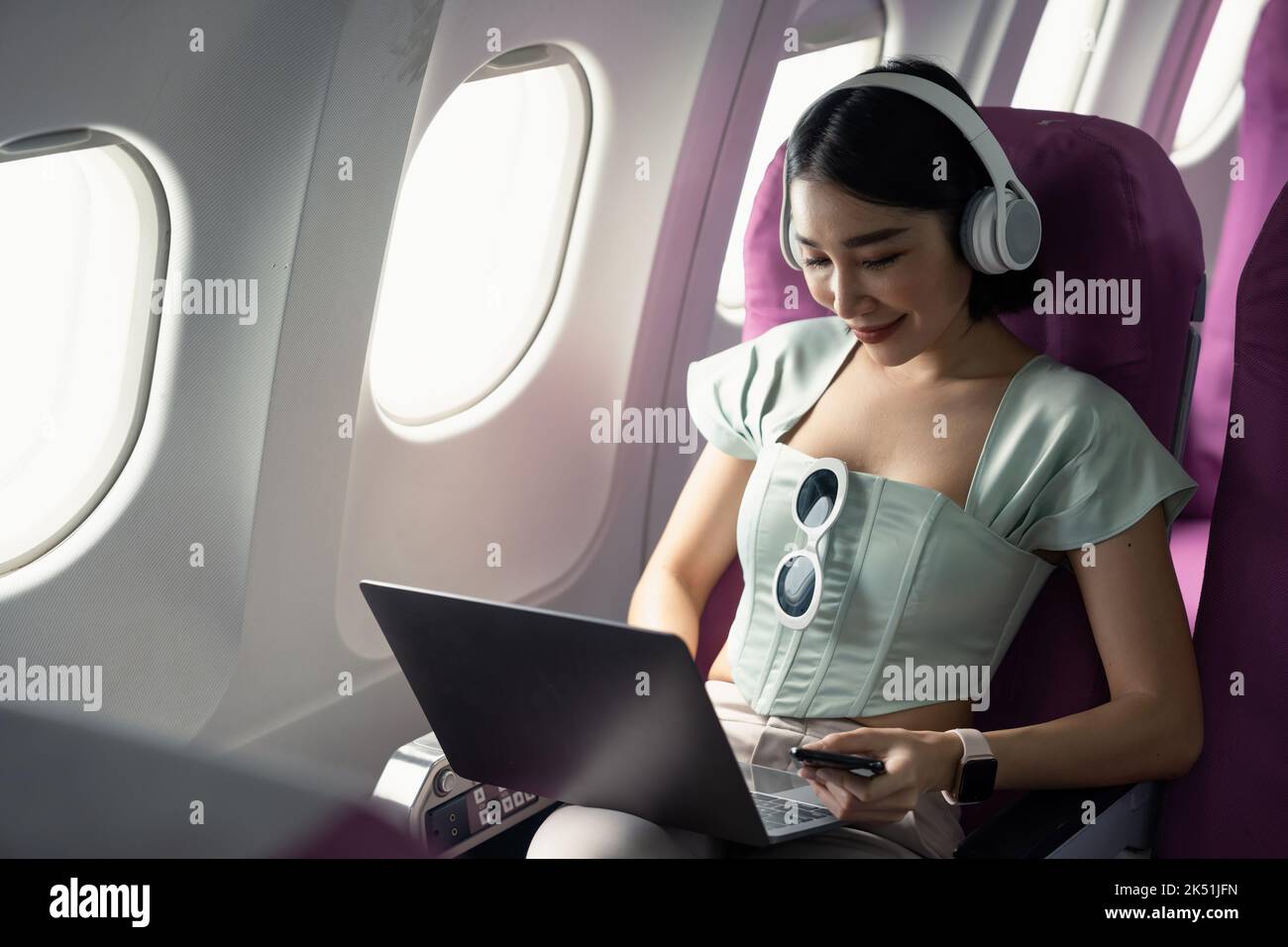 Plane passenger asian business woman working in airplane cabin during flight with laptop computer wifi listening to music with headphones Stock Photo
