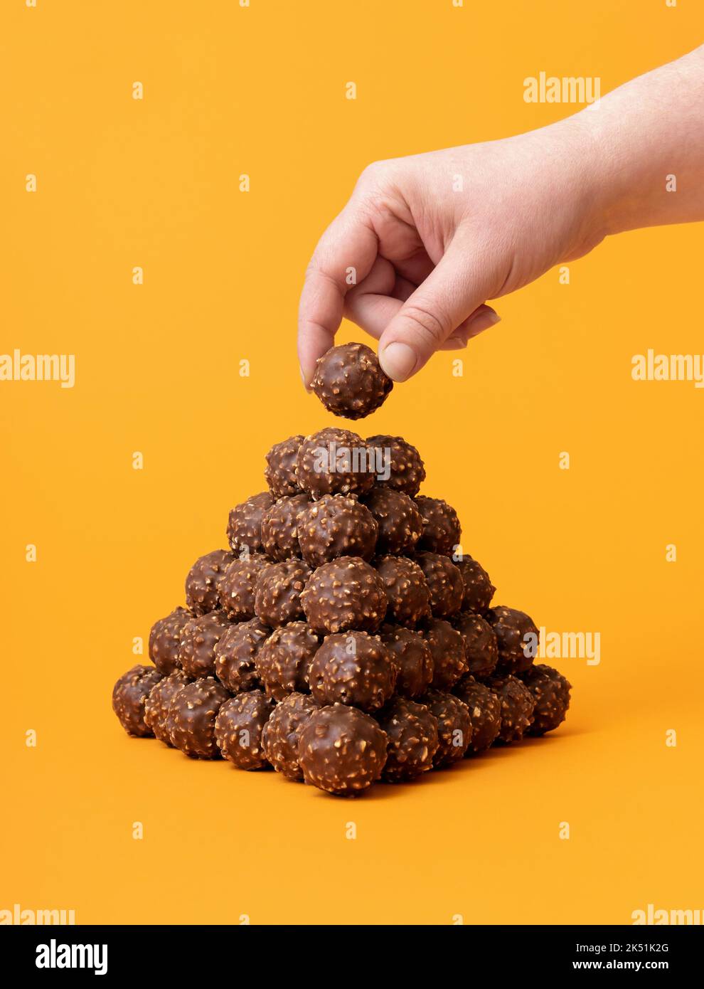 Woman's hand taking a chocolate truffle from a big pile. Chocolate candies tower on a vibrant orange background. Stock Photo