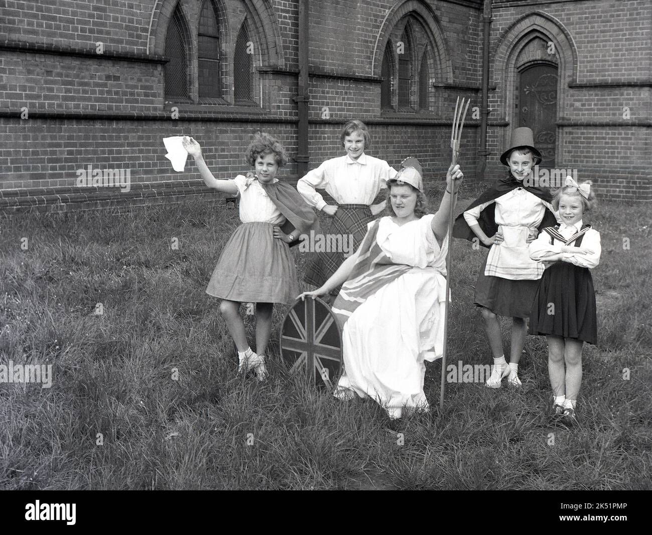 1961, historical, in the grounds of a local church, a group of girls taking part in the spring May Day celebrations pose together for a photo. In their parade costumes they stand around a teenage girl dressed as the goddess Britannia, wearing her Corinthian helmet, gown and with her trident and shield. Britannia, a helmeted female warrior, is an image dating back to the Roman province of Britain during the Roman Empire and was revived and became a popular symbolic figure in later periods of British history, particuarly in the age of the Empire. Stock Photo