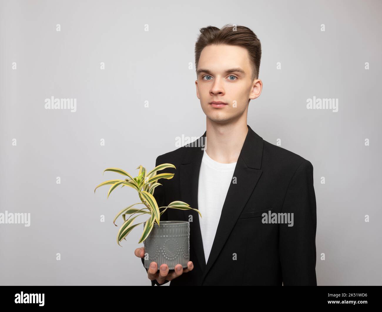 Portrait of elegant young man wearing black  suit holding potted house plant looking at camera. Studio shot on gray background Stock Photo