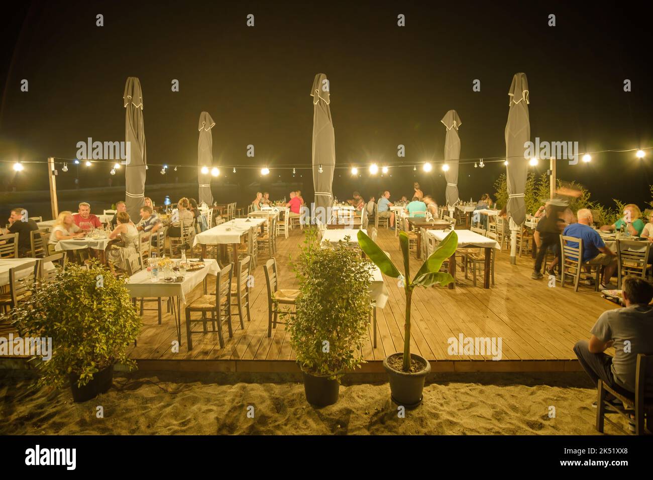 Nea Kallikrateia, Greece - August 31, 2022 : Greeks and tourists enjoying dinner outdoors at a cozy illuminated restaurant in front of the sea Stock Photo